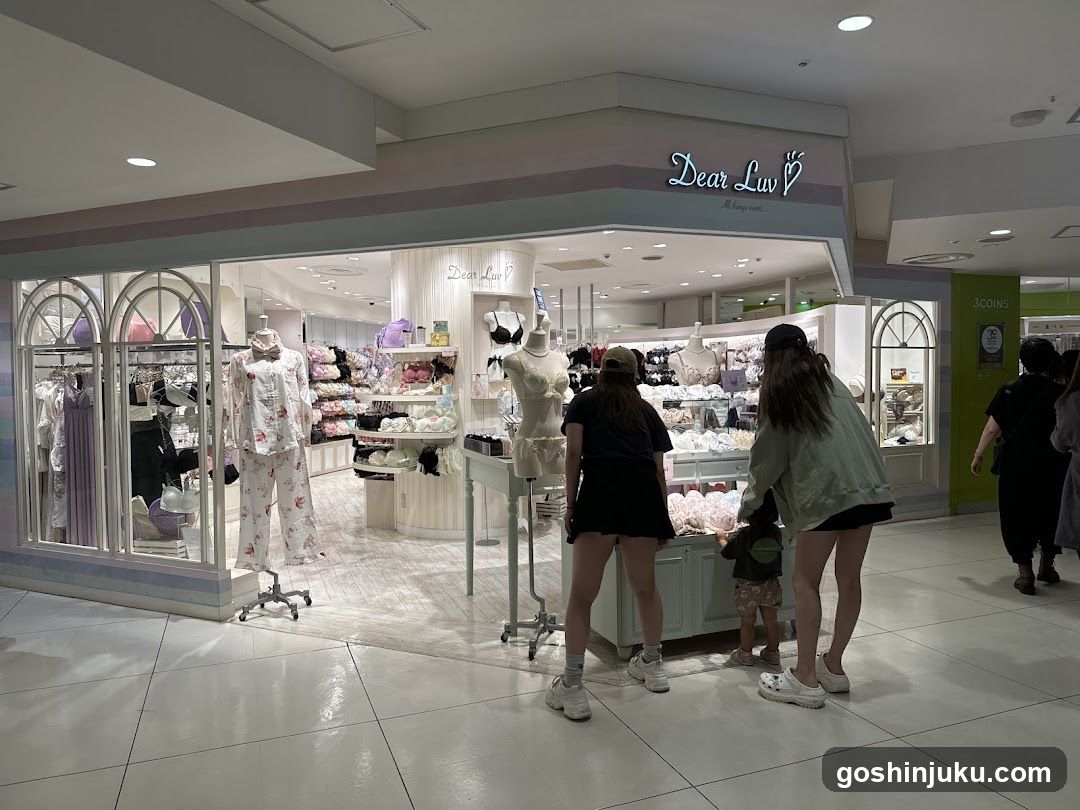 A brightly lit lingerie store with pastel-colored exterior and large glass windows displaying mannequins in pajamas and bras. The sign 'Dear Luv' is visible above the entrance. Inside, shelves are stocked with various lingerie items. Several customers browse near the entrance.