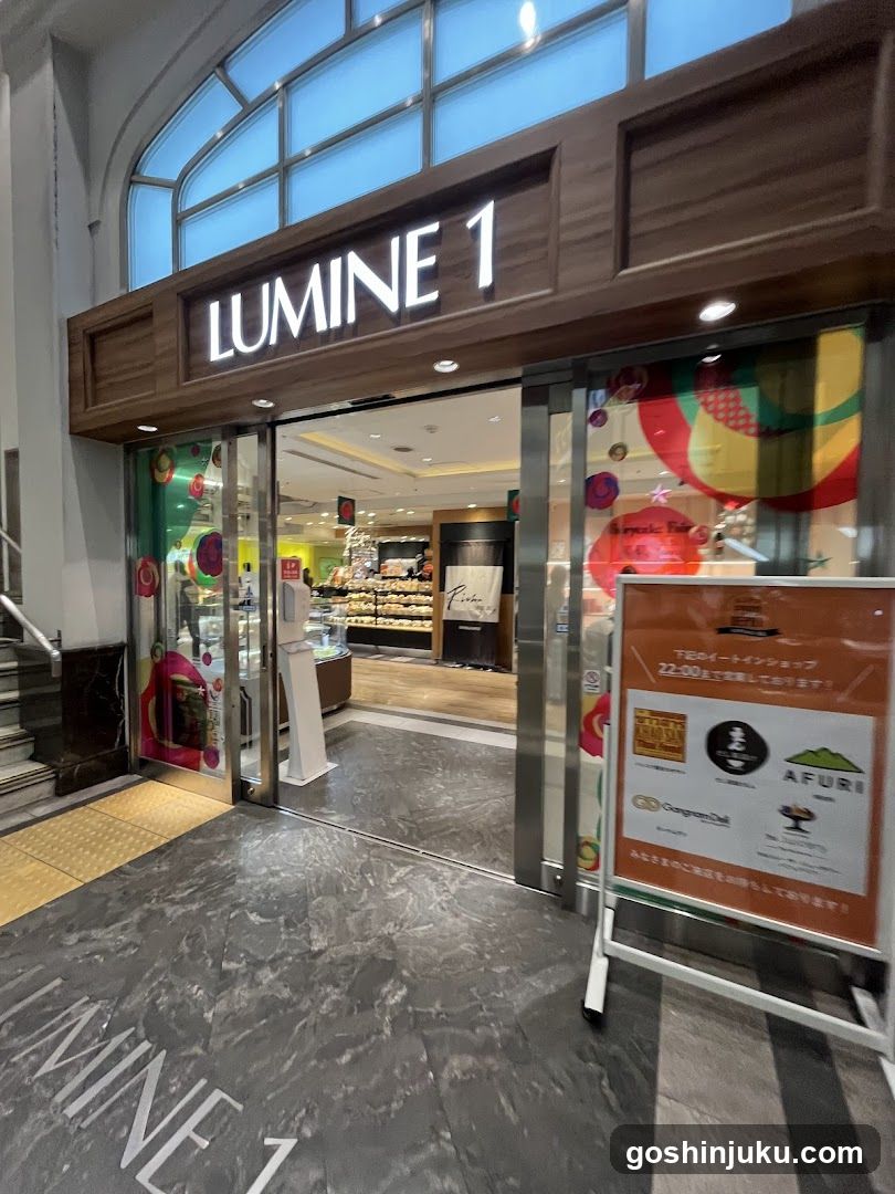 Photo of the physical exterior entrance of LUMINE 1, a shopping center in Japan. The storefront features glass doors with colorful circular decorations and signage above reading 'LUMINE 1'. A signboard outside advertises various food items including AFURI and other brands.