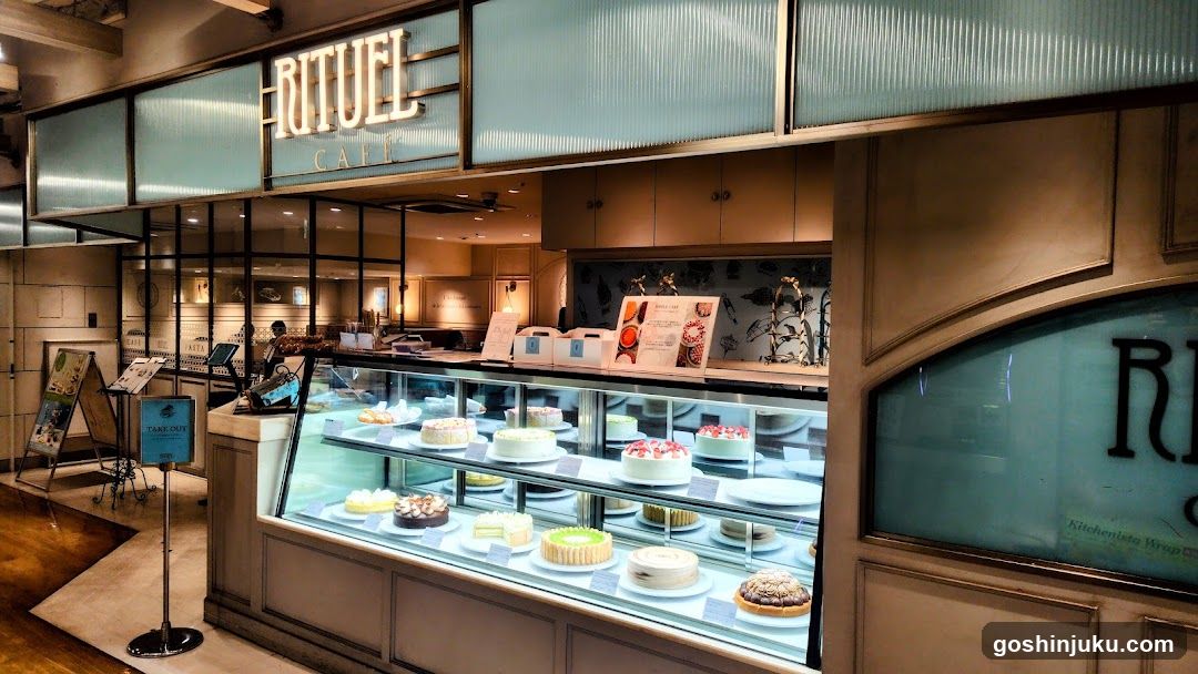 The image shows the storefront of Rituel Cafe, featuring a glass display case filled with various cakes and pastries. The shop sign is visible above the entrance, displaying 'RITUEL CAFE' in illuminated letters. Inside, there are additional displays and seating areas.