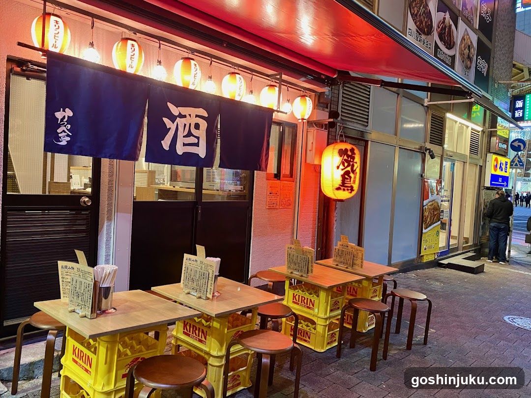 A nighttime street view of a Japanese izakaya with blue and white noren banners displaying kanji characters, including '酒' (sake). Yellow Kirin beer crates are repurposed as tables outdoors, each holding a menu board listing prices in yen. A glowing lantern hangs near the entrance, illuminating the sidewalk where pedestrians walk past adjacent shops.