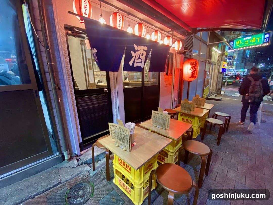 A small Japanese restaurant at night, featuring a red awning and hanging lanterns with the kanji '酒' (sake). Wooden tables on yellow crates line the sidewalk, with menus visible on each table. A person walks past in the background.