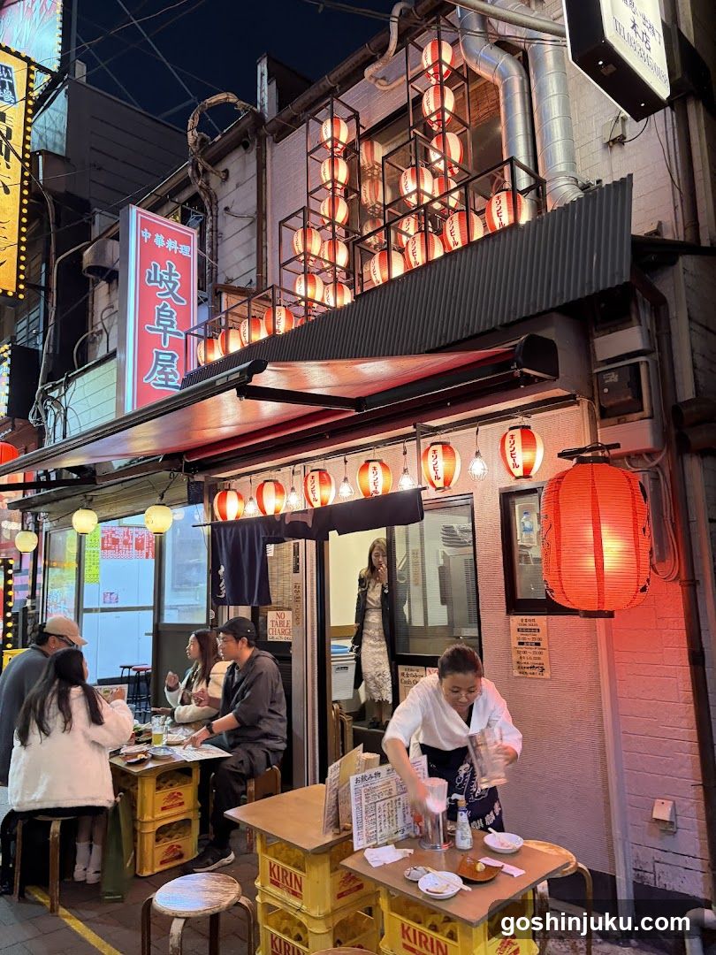 A traditional Japanese restaurant at night, featuring red lanterns and a sign with Japanese characters. Patrons are seated at tables made from yellow crates outside the entrance.