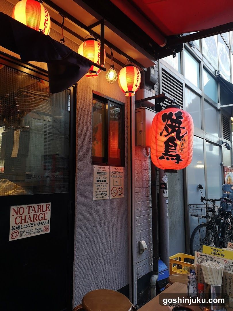 A shop front featuring a black door with 'NO TABLE CHARGE' signage, illuminated by red lanterns displaying Japanese characters. A menu board is visible on the wall next to the entrance.