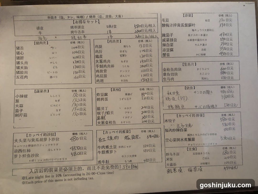 A printed menu board displaying various food items with prices in yen, organized into sections like 'Sets', 'Meat Dishes', 'Vegetables', and 'Salads'. The menu includes detailed descriptions of dishes such as pork belly rice, chicken curry, and vegetable salads.