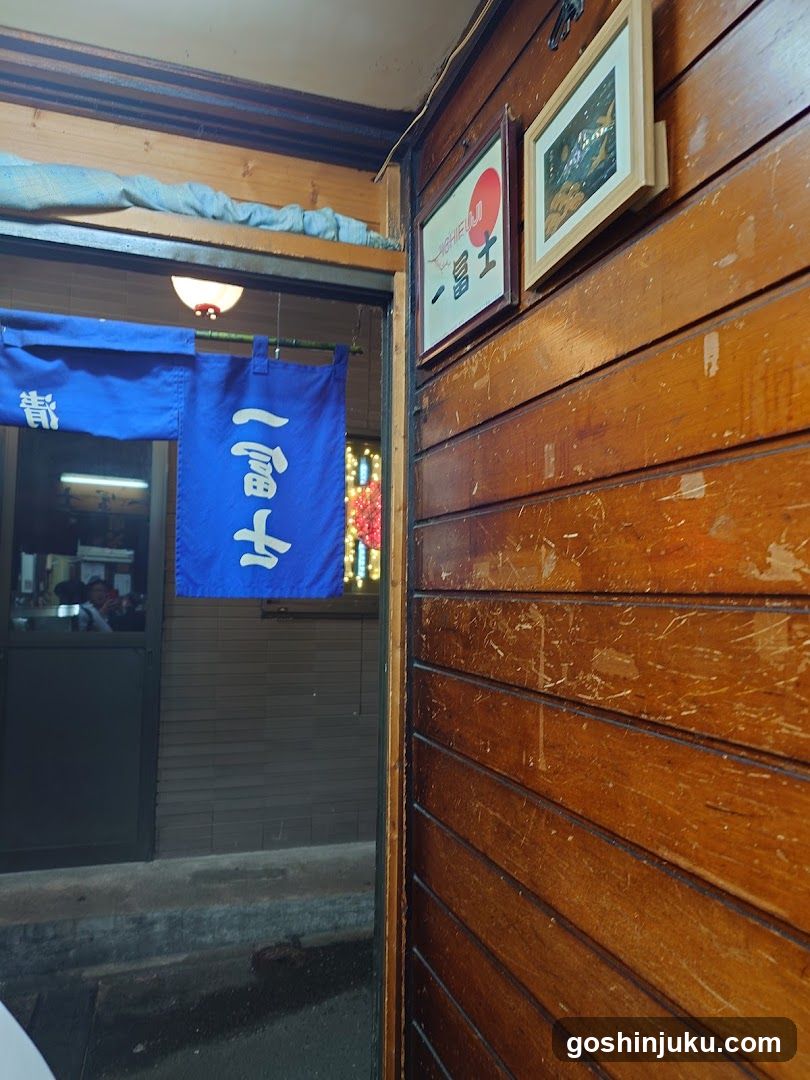 A wooden shop entrance with a blue noren (door curtain) displaying white Japanese characters. Above the door, framed artwork and signs are mounted on the wall. The interior is dimly lit, suggesting a cozy, traditional atmosphere.