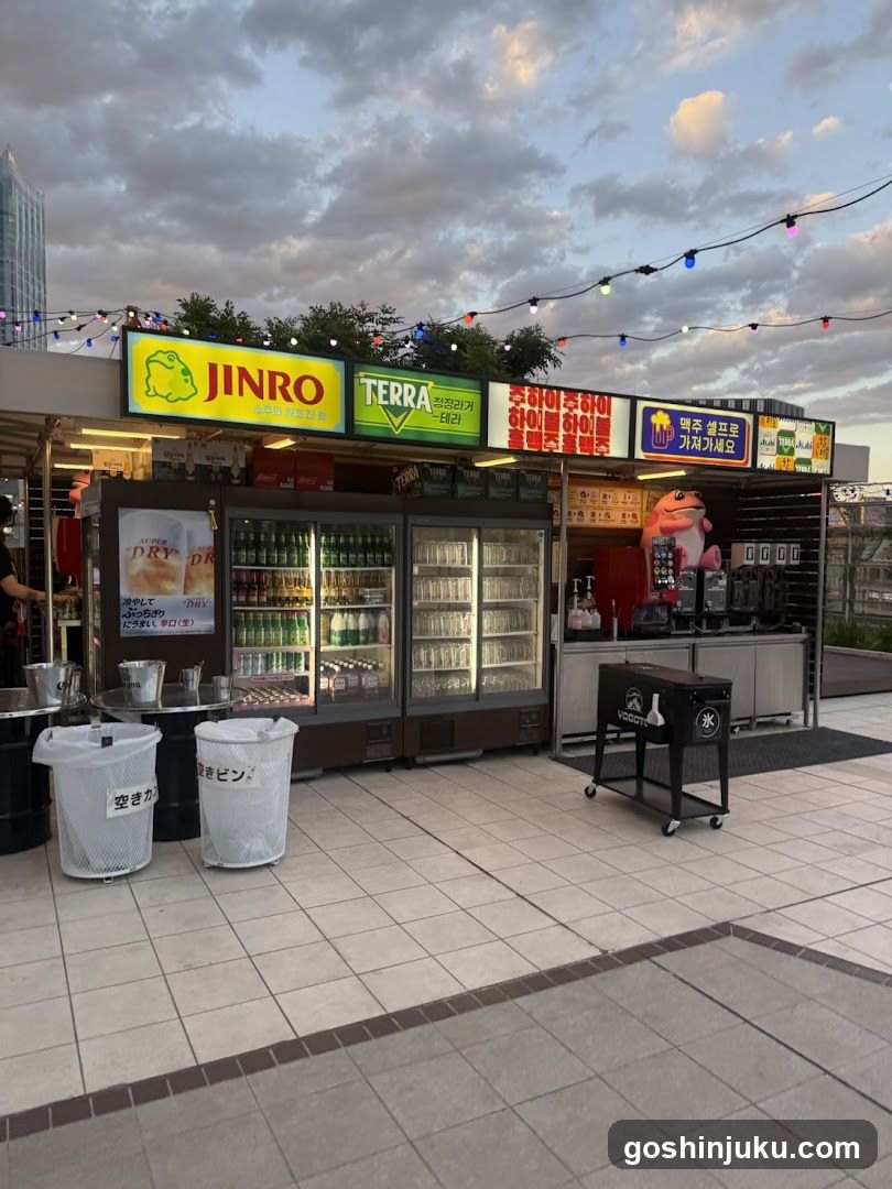 Outdoor food and drink stalls with visible signage for Jinro (green bottle logo) and TERRA (green square logo). Refrigerated display cases filled with bottled drinks, ice buckets, trash bins labeled in Japanese, and a black cart with branding. String lights overhead against cloudy sky.