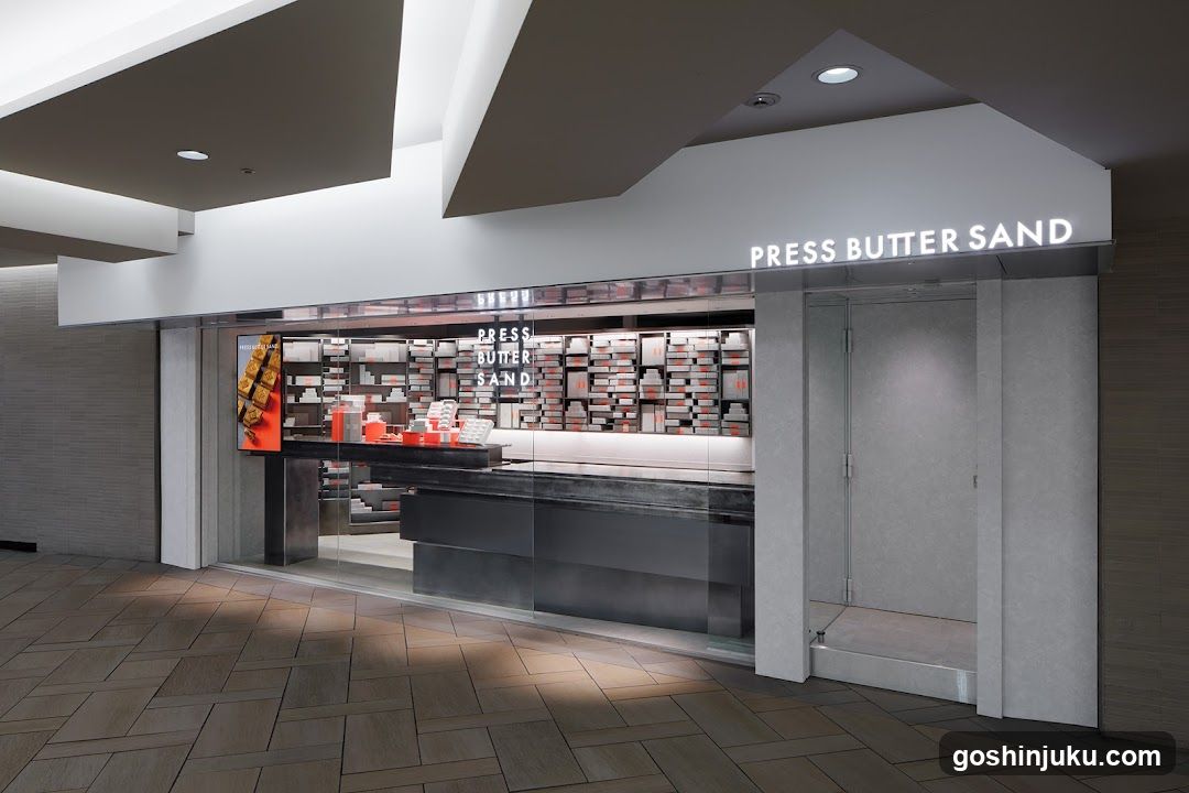 Modern storefront with illuminated signage reading 'PRESS BUTTER SAND' above entrance. Glass display window shows neatly arranged products, including red and white boxes. Interior features dark counter and shelving units displaying merchandise. Entrance door is closed with metallic frame.