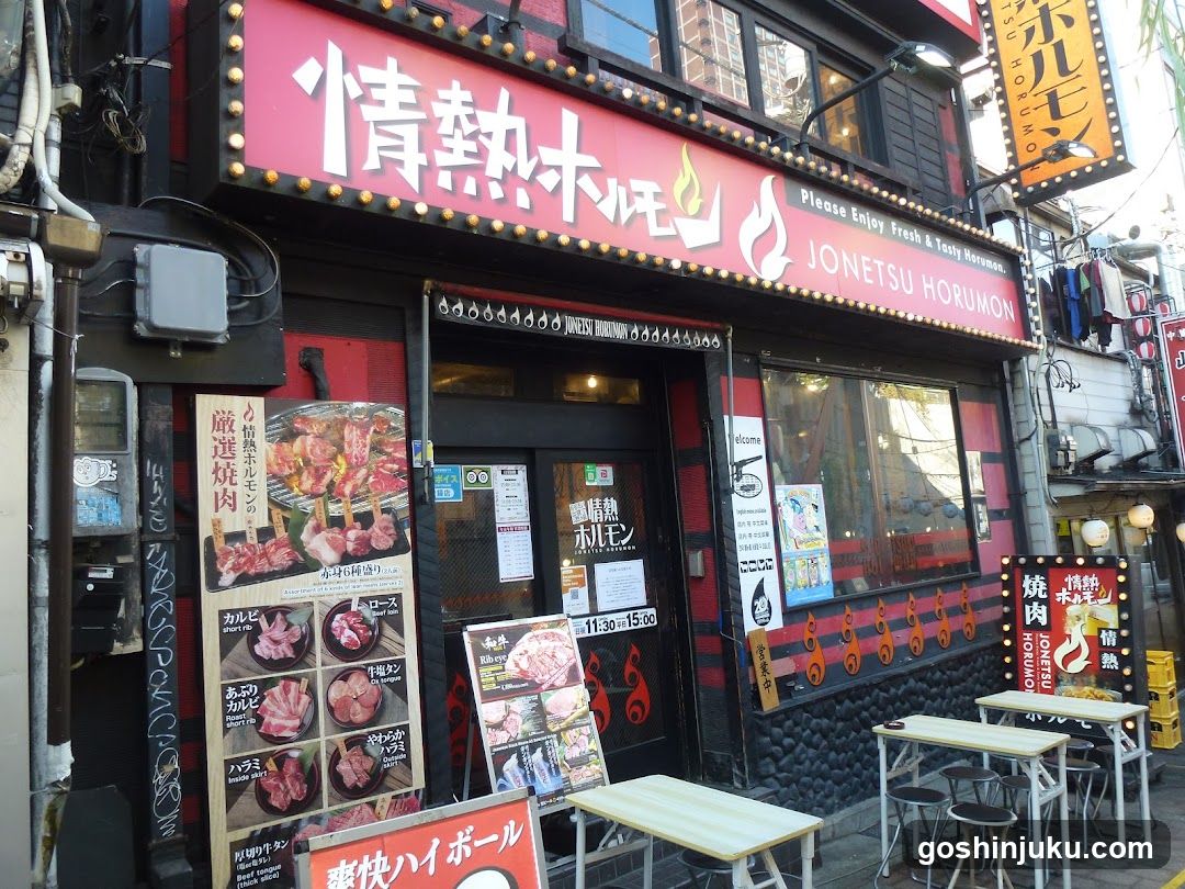 A street-level view of a yakitori and yakiniku restaurant called Jonetsu Horumon, featuring red signage with Japanese characters and English text 'Please Enjoy Fresh & Tasty Yakiniku'. The entrance has glass doors with safety notices and posters. Outside tables are set up on the sidewalk. A vertical menu board displays grilled meat options like beef ribs, pork belly, and chicken wings. Additional signs advertise special promotions and operating hours from 11:30 to 15:00.