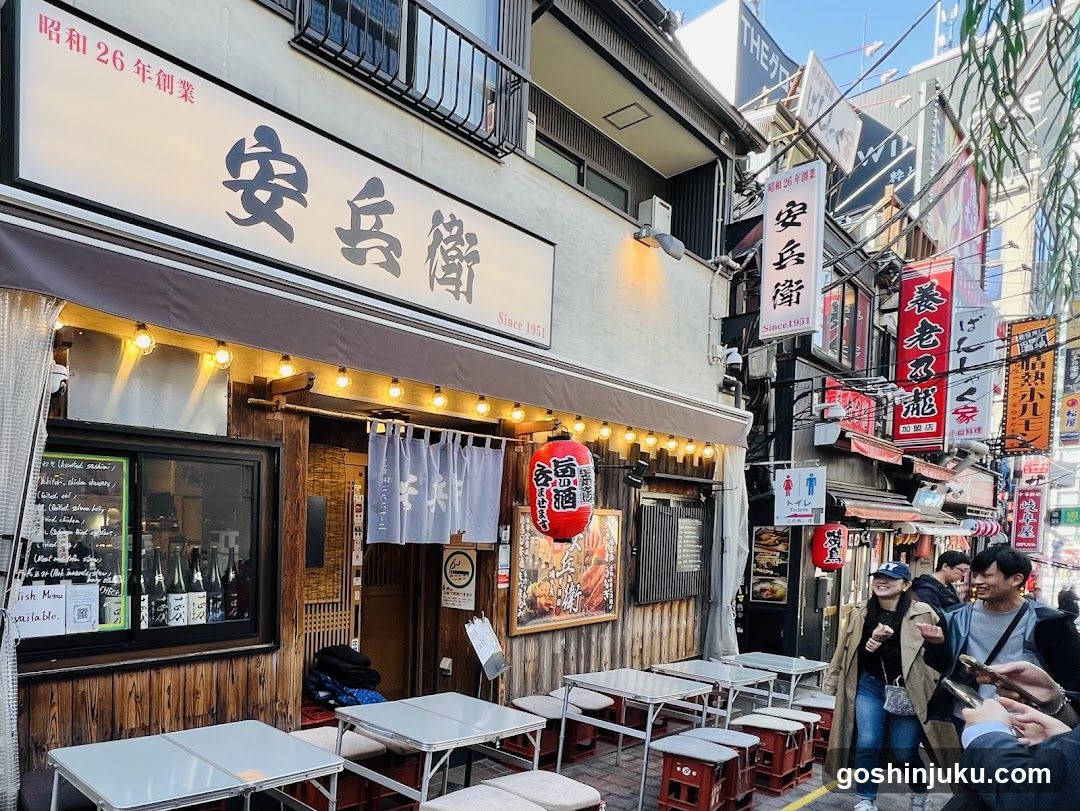 A traditional Japanese shop front with a white facade and wooden accents, featuring large black kanji characters on the signboard reading '安兵衛' (Anboshu). The entrance is adorned with hanging paper lanterns and string lights, creating a warm ambiance. Outside seating includes small tables and stools arranged along the sidewalk. A red lantern hangs near the door, adding to the cultural aesthetic. Surrounding buildings display various signs, indicating a bustling street scene.
