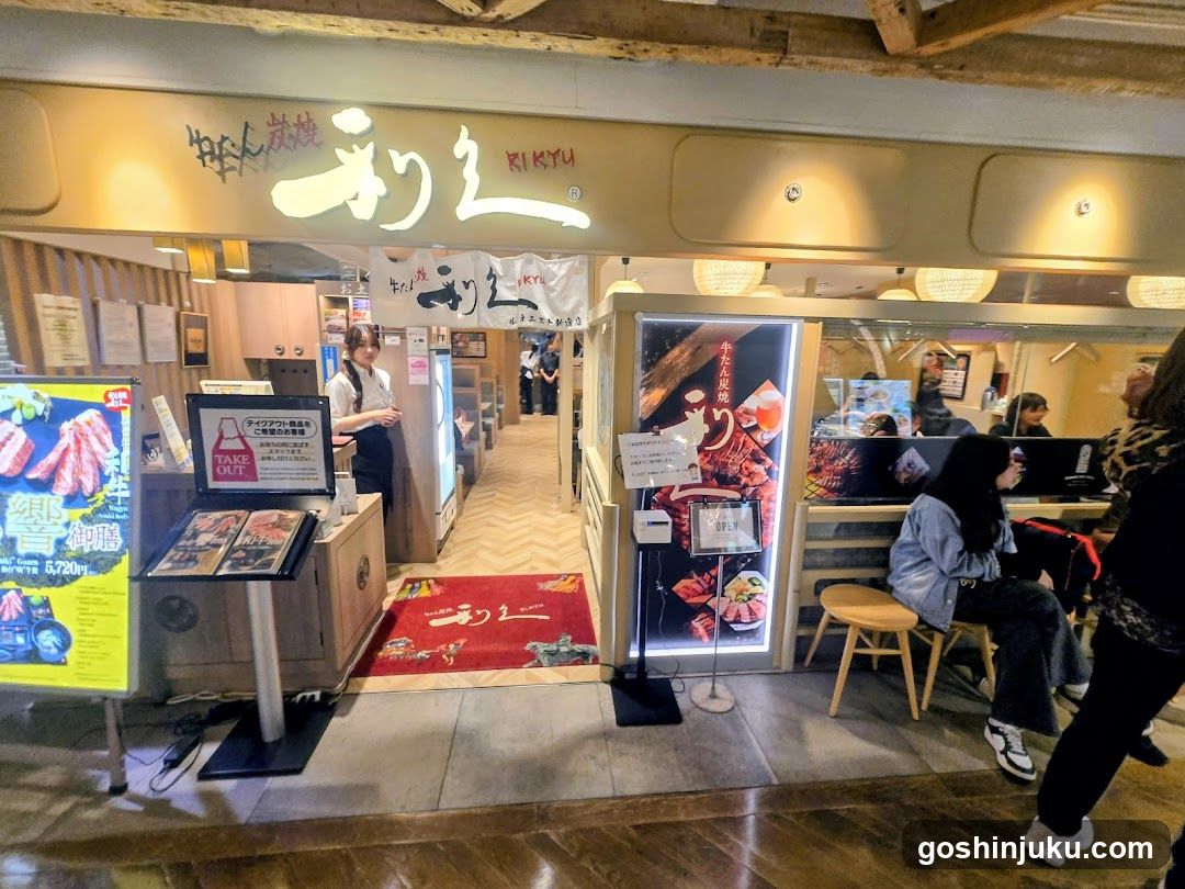 The image shows the entrance of a Japanese restaurant named Rikyū, with illuminated signage above the door. A red welcome mat is placed at the entryway. Inside, staff members are visible near the counter and kitchen area. Outside, customers sit on wooden stools near the entrance. Promotional posters and menu boards display food items and prices.