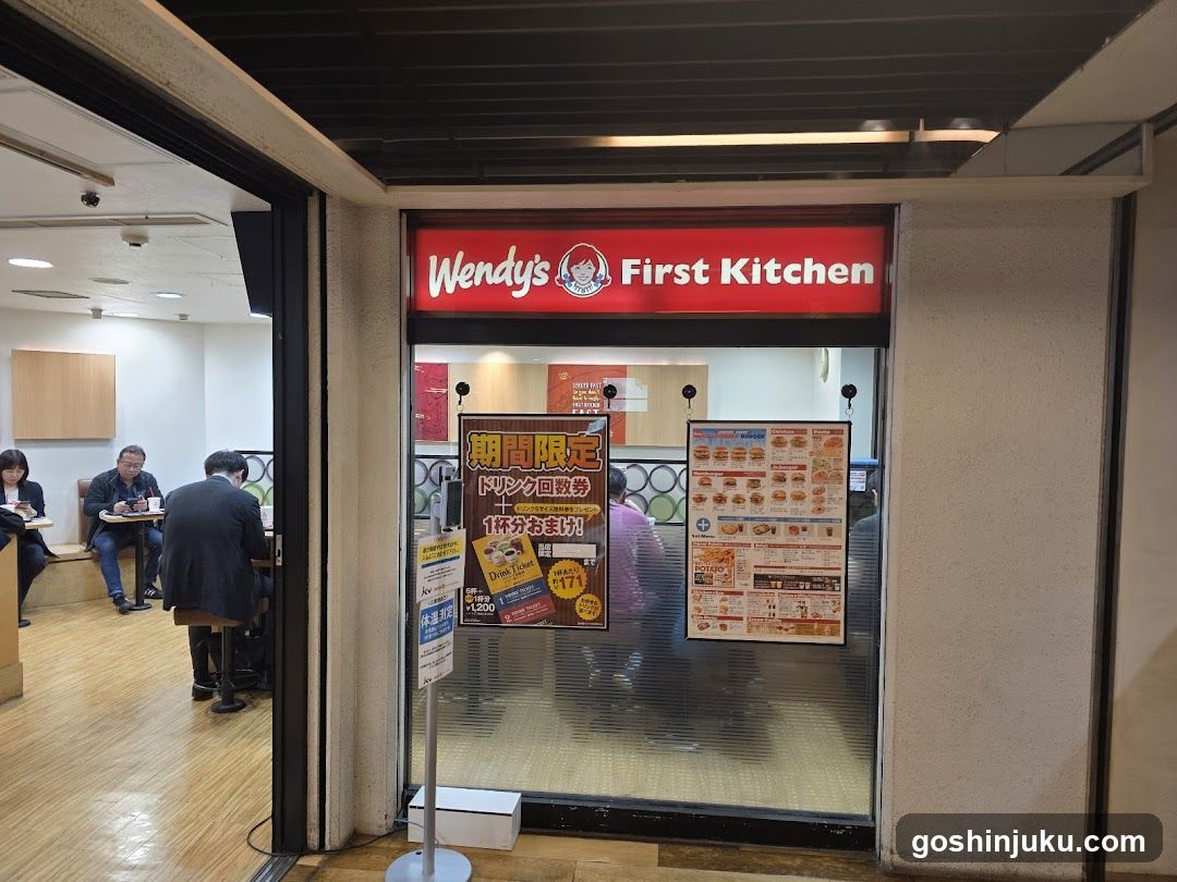 Photo of the physical storefront entrance to Wendy's First Kitchen, featuring a red illuminated sign with the brand logo and name above glass doors. Inside, customers are seated at tables in a casual dining area.