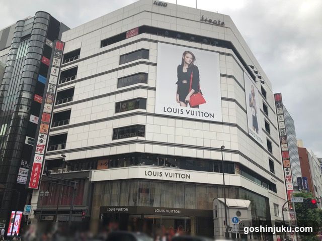 A multi-story building with a prominent Louis Vuitton storefront at the ground level, featuring large glass windows and signage displaying 'LOUIS VUITTON' above the entrance. A large billboard on the side of the building showcases a model wearing a black outfit with a red handbag, also branded with 'LOUIS VUITTON'. The surrounding area includes other commercial buildings with vertical signs in Japanese characters.