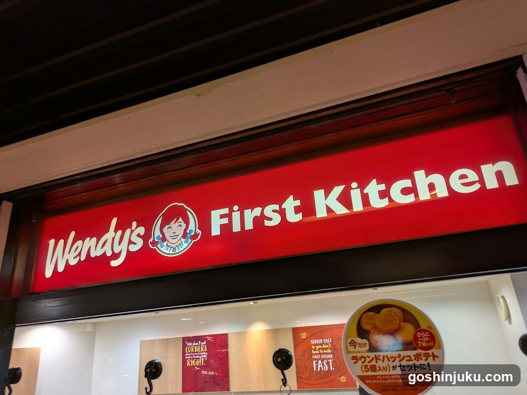 A brightly lit storefront featuring a red sign with 'Wendy's' logo and the text 'First Kitchen'. Below, there are promotional posters advertising food items like 'Round Hash Potatoes', with Japanese text indicating special sets.