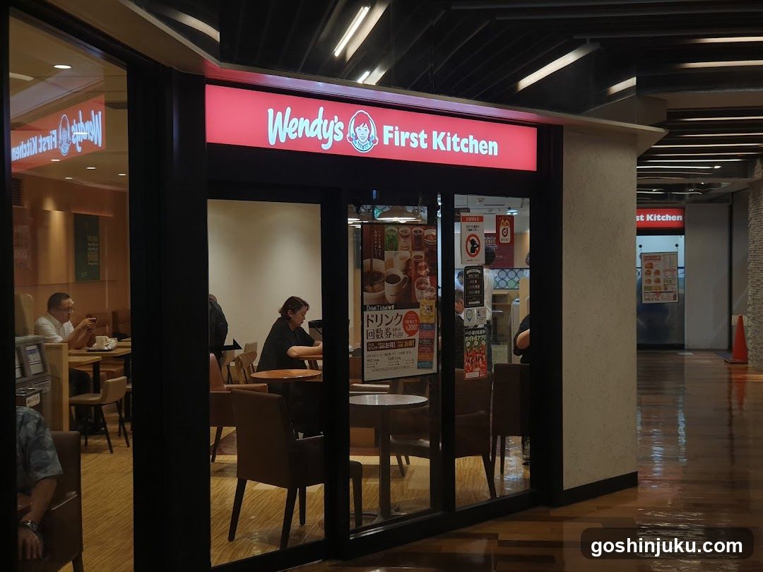 A Wendy's First Kitchen storefront with a red illuminated sign displaying the brand name and logo. The entrance features glass doors and windows, revealing customers seated at tables inside. Posters and advertisements are visible on the window, including one promoting a special offer.