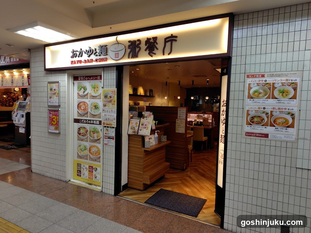 The image shows the storefront of a noodle shop with white tiled walls and wooden accents. The sign above reads 'Oka-yu & Noodle' in Japanese characters, along with an illustration of a bowl of noodles. Inside, there is a counter area with menus displayed on the wall. Posters featuring various noodle dishes are visible both inside and outside the store.