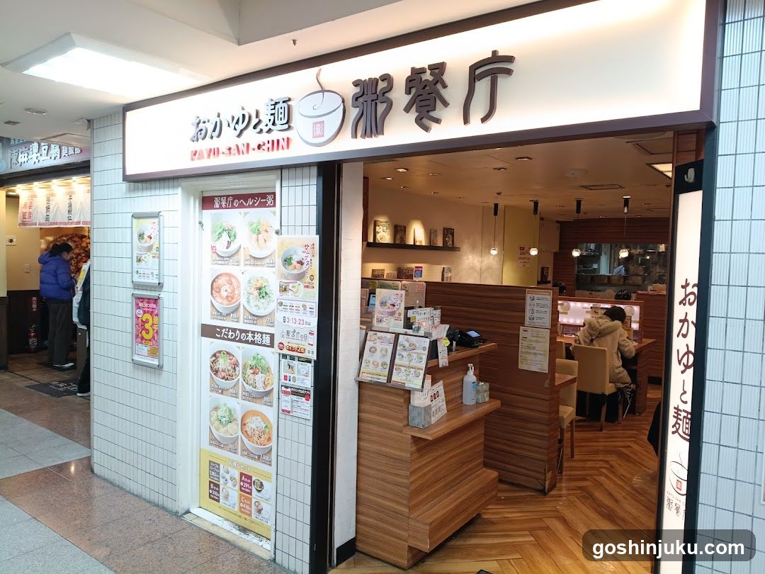 The image shows the exterior entrance of a restaurant, featuring prominent signage above the door with Japanese characters and English text 'OKAYU & Noodles'. The storefront includes a tiled facade and wooden interior elements visible through the open doorway. A large menu board is displayed on the left side, showcasing various noodle dishes with prices in yen. Inside, customers are seated at tables, and the warm lighting creates an inviting atmosphere.