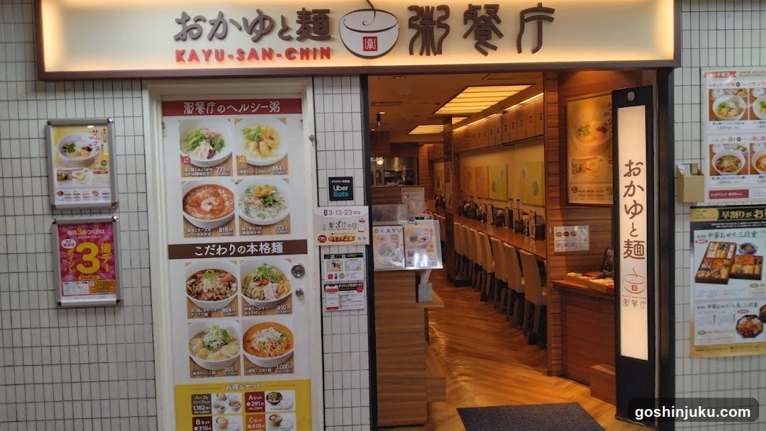 The image shows the entrance of a noodle shop with a signboard displaying 'Okayu-to Noodles' in Japanese and English. The door is open, revealing the interior seating area with wooden tables and chairs. There are posters on the walls inside and outside the shop.