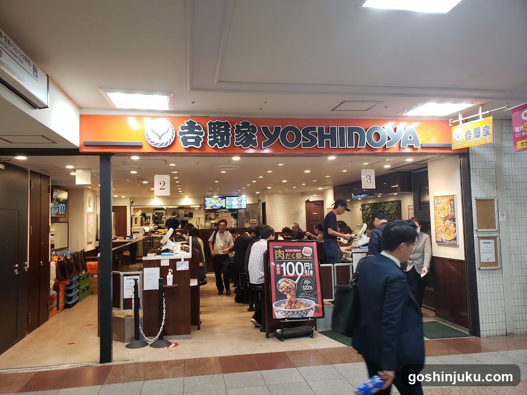 The image shows the entrance of a Japanese restaurant named Yoshinoya, with its name displayed prominently in both Japanese and English on an orange sign above the door. The store is bustling with customers waiting inside, some standing near the counter while others are seated at tables. A large promotional poster advertises a special offer for 100 yen items. Inside, there are multiple sections marked by numbers, indicating different areas or services offered by the establishment.