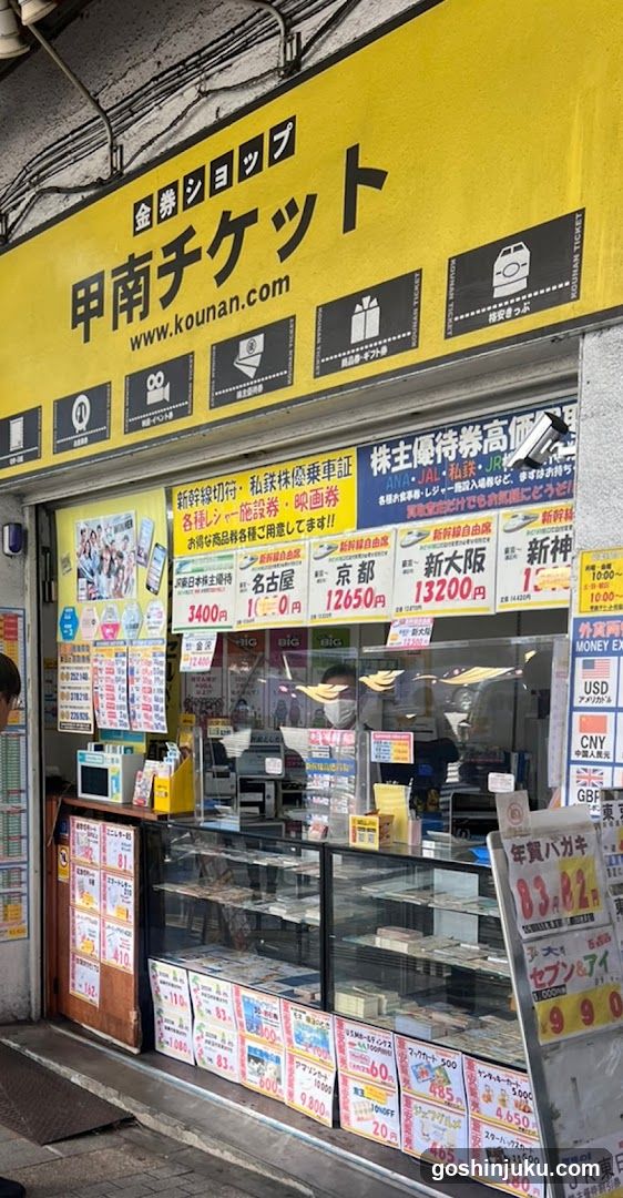 A yellow storefront with signage reading '甲南チケット' (Kouan Ticket) and website www.kounan.com. The shop sells tickets, gift cards, and travel vouchers. Visible items include price lists on the window, a glass display case filled with merchandise, and posters advertising train tickets to destinations like Kyoto, Osaka, and Shinagawa.