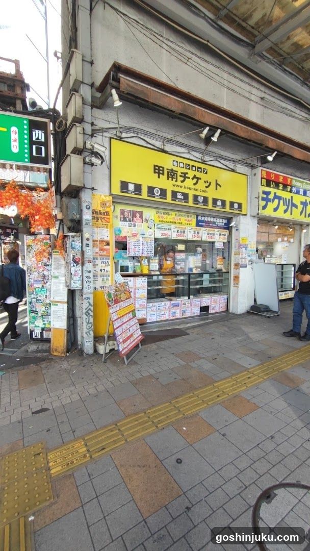 A street-level view of a ticket shop facade with yellow signage reading '甲南チケット' (Kinan Tickets). The storefront features glass windows displaying various posters and advertisements, including concert tickets and event listings. A person is standing near the entrance, and another individual is visible to the left. The building has exposed pipes and wires above the shop, typical of urban architecture.
