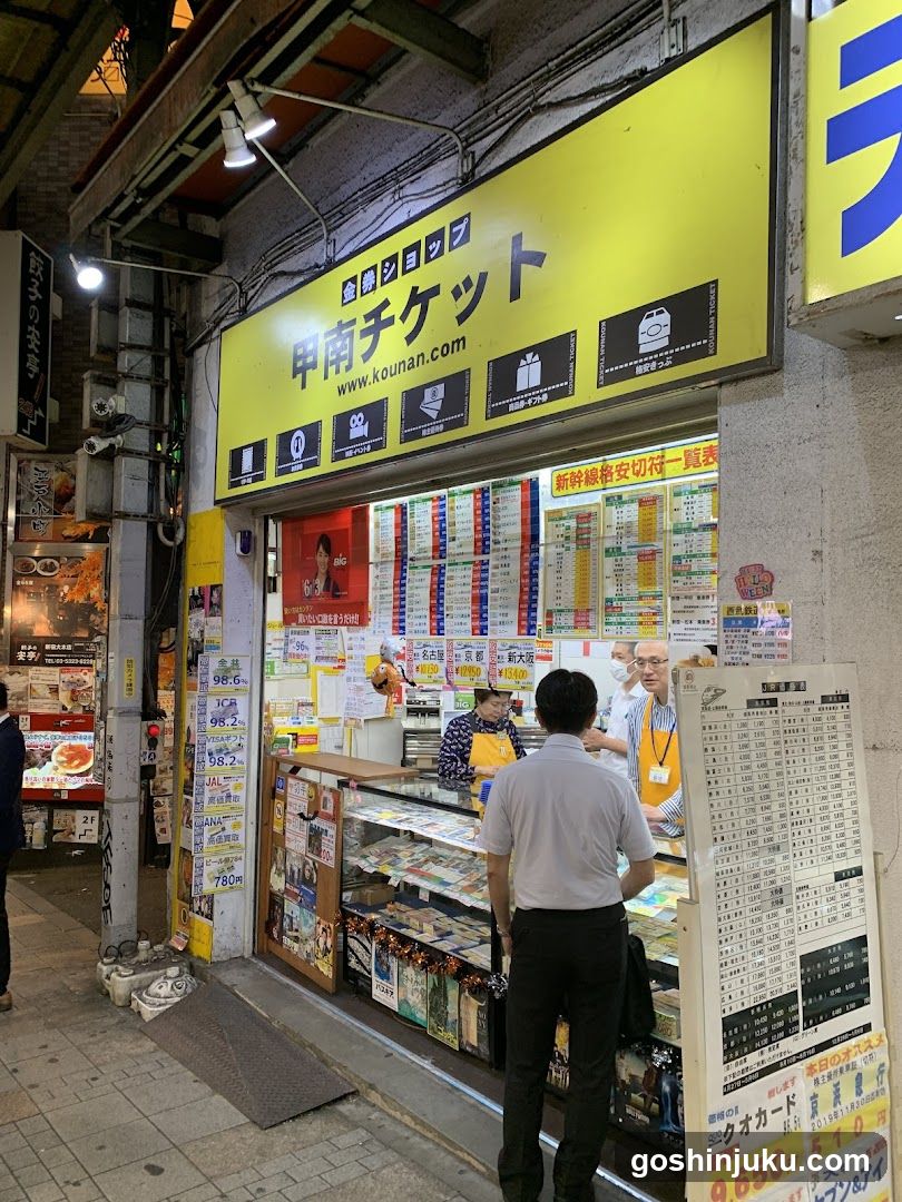 A brightly lit ticket shop with a yellow sign reading '甲南チケット' and website 'www.kounan.com'. The storefront displays numerous schedules, posters, and price lists inside the glass window. A man in a white shirt stands at the counter facing two staff members wearing orange aprons. Outside to the right is a large menu board listing train tickets and prices.