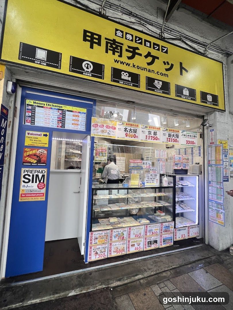 A shop front with a yellow sign reading '甲南チケット' (Kounan Ticket) and website 'www.kounan.com'. The entrance has blue doors, one open revealing the interior. Inside, there are glass display cases filled with tickets or vouchers. Posters advertise prepaid SIM cards and currency exchange rates. A staff member is visible behind the counter.