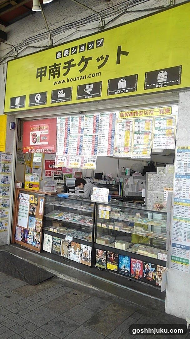 A shop front with a bright yellow sign displaying '甲南チケット' (Kouan Ticket) and the website www.kounan.com. The storefront features glass display cases filled with various items, likely tickets or merchandise. Inside, there are numerous posters and advertisements on the walls, including one mentioning '95%'. A person is visible inside the shop, possibly attending to tasks.