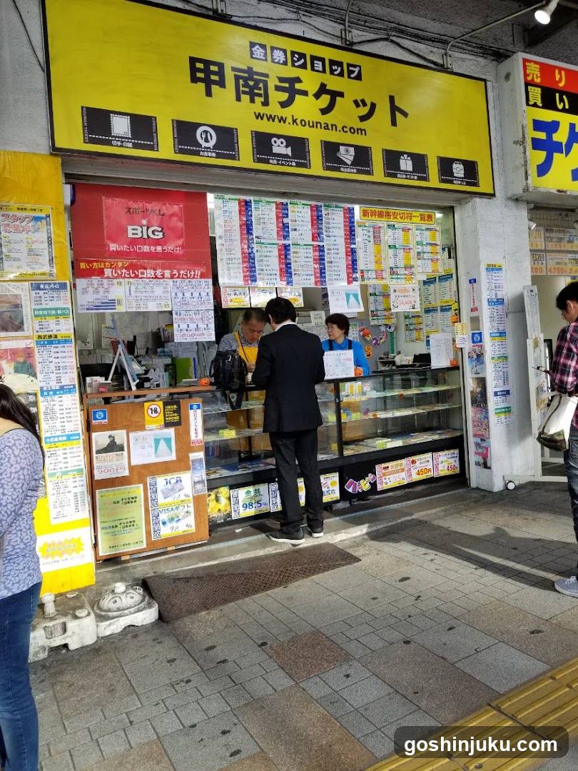 A street-level view of a ticket shop with a yellow sign reading '甲南チケット' (Kounan Ticket). The storefront features glass windows displaying various schedules and posters, including train timetables and event listings. A customer in a suit is standing at the counter interacting with staff. Posters advertise services like 'BIG' and other local events.
