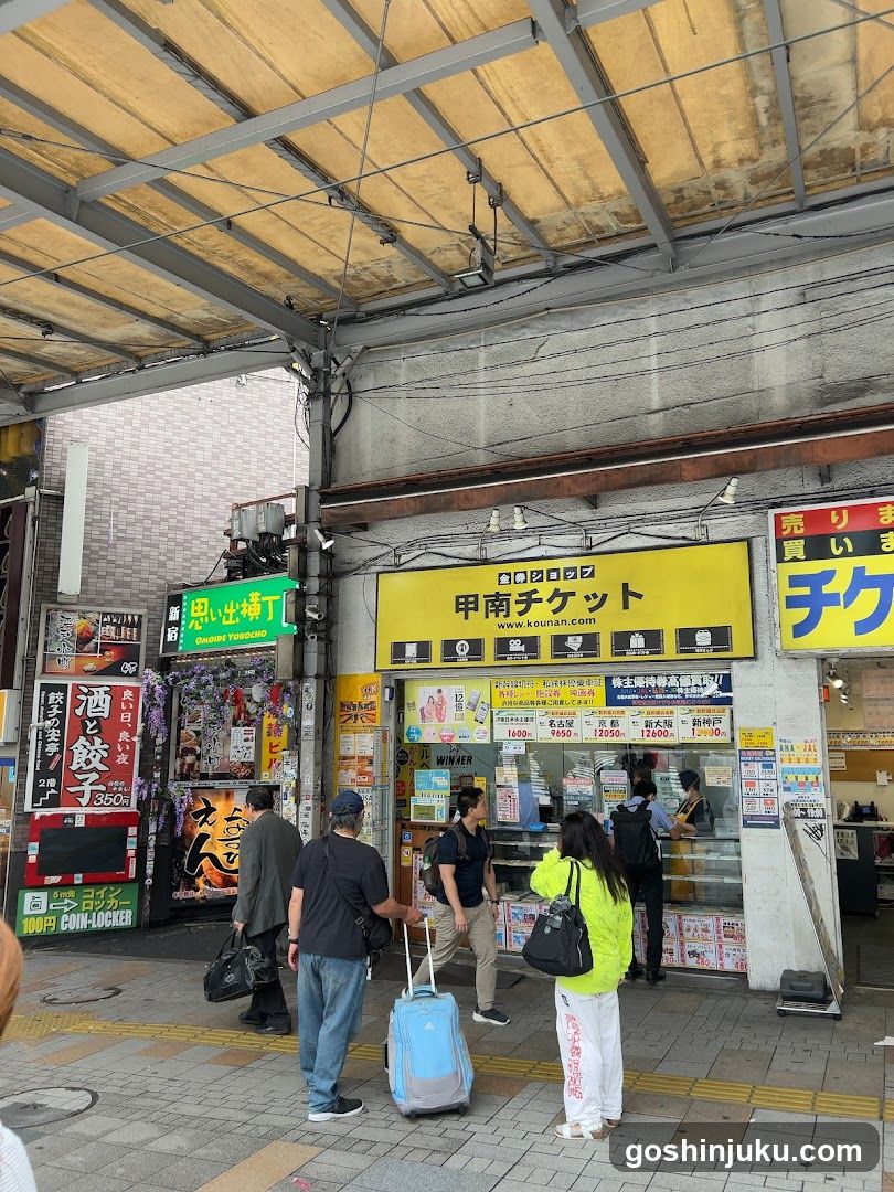 A street-level view of a ticket shop with yellow signage reading '甲南チケット' (Kominato Ticket). The storefront features glass windows displaying various posters and advertisements. People are standing outside, some holding luggage, suggesting travel or transit activity. Adjacent shops include a convenience store and other local businesses.