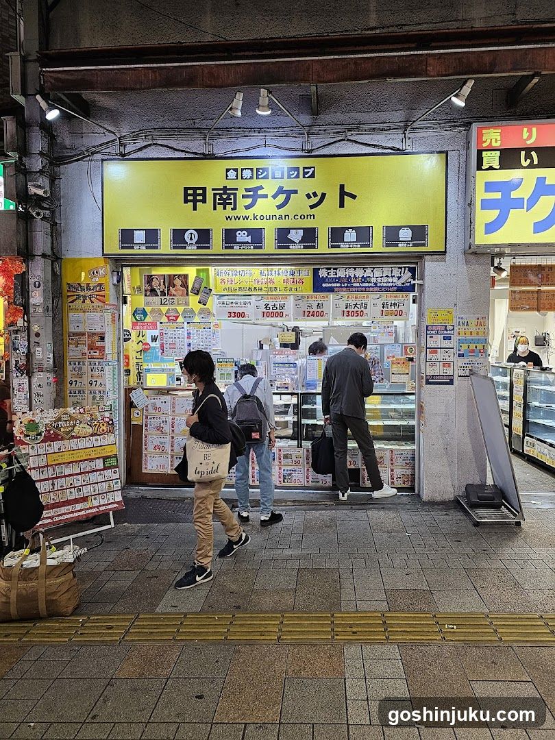 A street-level view of a ticket shop with bright yellow signage reading '甲南チケット' (Kounan Ticket) and website www.kounan.com. The storefront is filled with colorful posters, price lists, and promotional materials. Several people are browsing or entering the shop. A large display case is visible to the right.