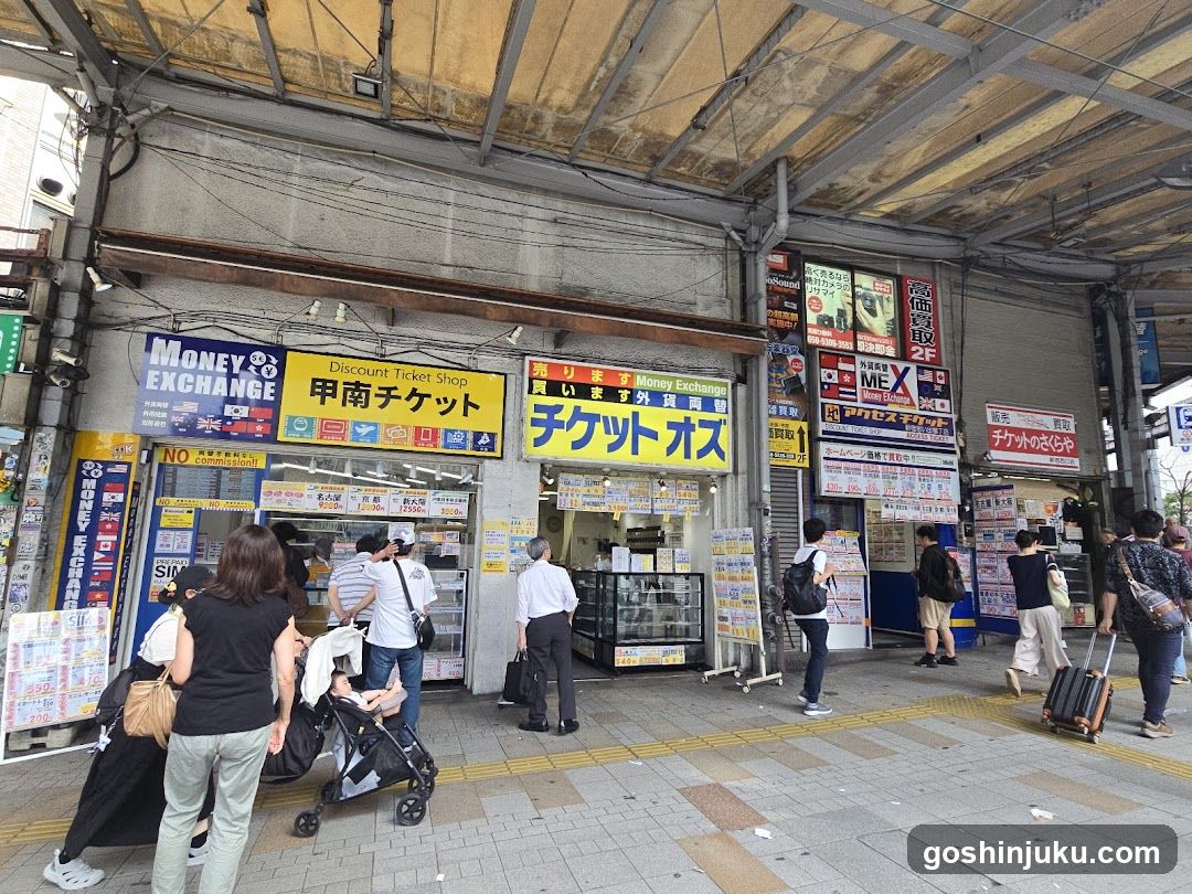 A street-level view of a row of small shops under an awning, featuring multiple signs including 'Money Exchange', 'Discount Ticket Shop', and 'チケット オズ'. People are walking by with luggage.