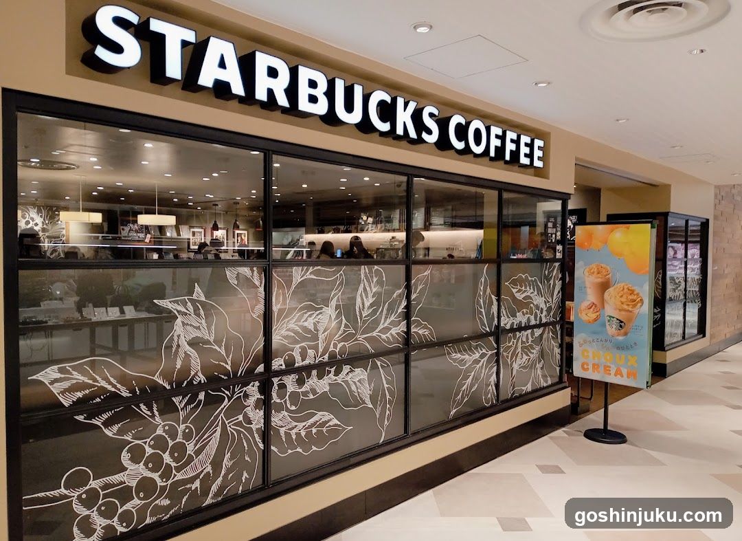 A Starbucks Coffee storefront with large glass windows featuring white botanical illustrations of coffee leaves and berries. The sign 'STARBUCKS COFFEE' is prominently displayed above the entrance in bold, illuminated letters. A promotional standee outside advertises a drink called 'Choco Cream'.