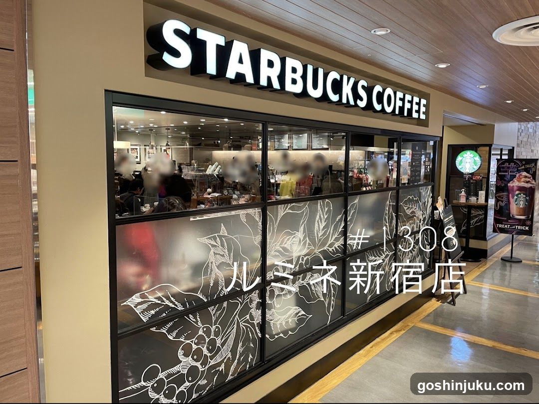 A Starbucks Coffee storefront with illuminated signage reading 'STARBUCKS COFFEE' above a large glass window featuring frosted panels with coffee bean illustrations and Japanese text. Inside, customers are seated at tables, and barista activity is visible through the glass.