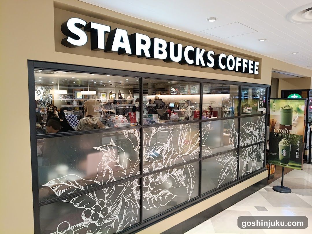 A Starbucks Coffee storefront with large glass windows featuring white botanical illustrations of coffee leaves and berries. The sign 'STARBUCKS COFFEE' is prominently displayed above the entrance in bold, illuminated letters. Inside, customers are seated at tables, and staff members are visible behind the counter. A promotional poster for 'Yokai Matcha' stands outside near the entrance.