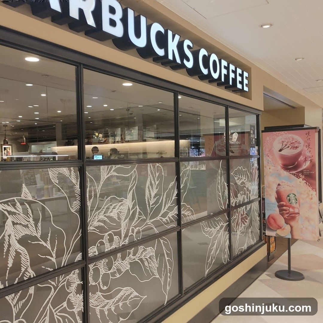 Exterior view of a Starbucks Coffee store with illuminated signage reading 'STARBUCKS COFFEE' above large glass windows featuring white botanical leaf patterns. A promotional poster stands outside displaying coffee cups and seasonal imagery.