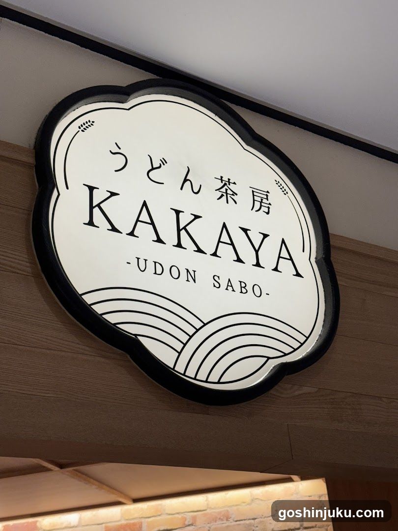 A close-up photo of a shop sign mounted on the exterior wall above an entrance. The sign is oval-shaped with a black border and white background, featuring Japanese characters (うどん茶房), the English name 'KAKAYA', and '-UDON SABO-' below it. Decorative wave patterns are at the bottom. Below the sign, part of a brick facade and wooden trim are visible.