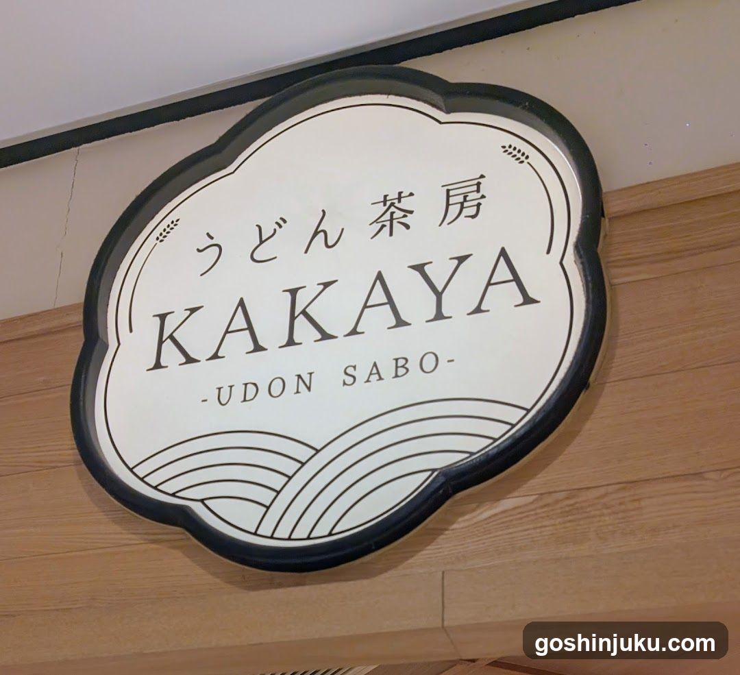 A close-up photo of a shop sign mounted on a wooden wall. The sign is oval-shaped with a black border and white background. It features Japanese characters 'うどん茶房' (Udon Chabou) at the top, followed by the English name 'KAKAYA', and '-UDON SABO-' below it. Decorative wave-like lines are visible at the bottom of the sign.