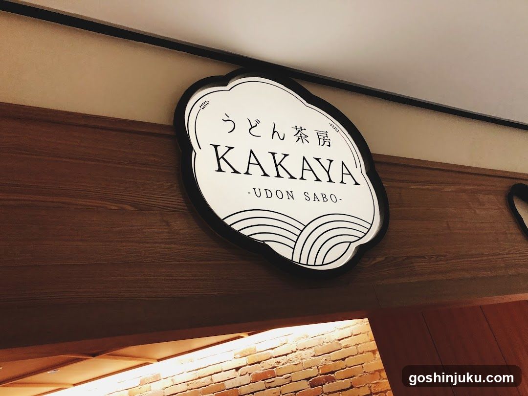 A close-up photo of a shop sign mounted on wooden wall paneling above an entrance. The sign is oval-shaped with black border and white background, displaying Japanese characters 'うどん茶房' (Udon Cha-ya), the English name 'KAKAYA', and '-UDON SABO-' below it. Decorative curved lines resembling noodles or waves are at the bottom. Below the sign, part of a brick wall and warm lighting from inside the shop are visible.