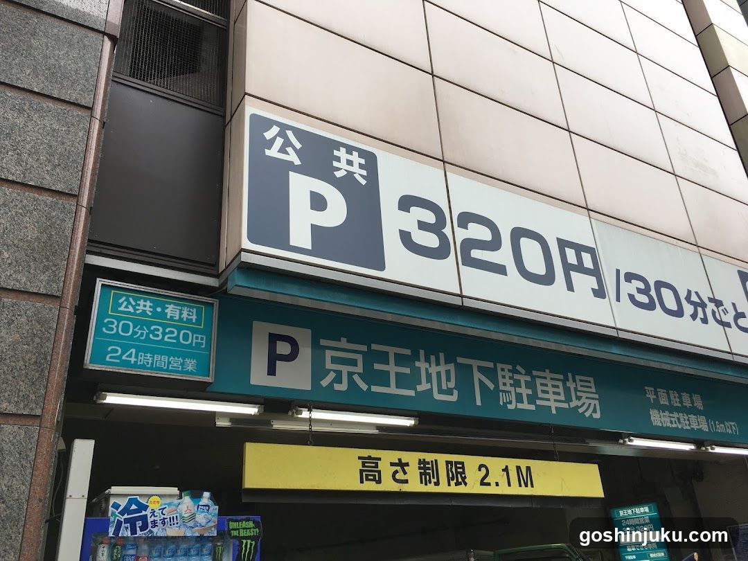 Exterior view of a parking garage entrance with signage indicating public parking, pricing (320 yen per 30 minutes), and height restriction (2.1M). The building facade features tiled walls and illuminated signs in Japanese.