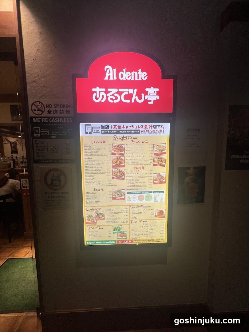 A photo of the physical exterior entrance to a restaurant named Al dente (あるでん亭). A large illuminated red sign with white text is mounted on the wall above the door. To the left, there are several signs including 'NO SMOKING' and 'WE'RE CASHLESS'. Below the main sign hangs a menu board displaying various food items.