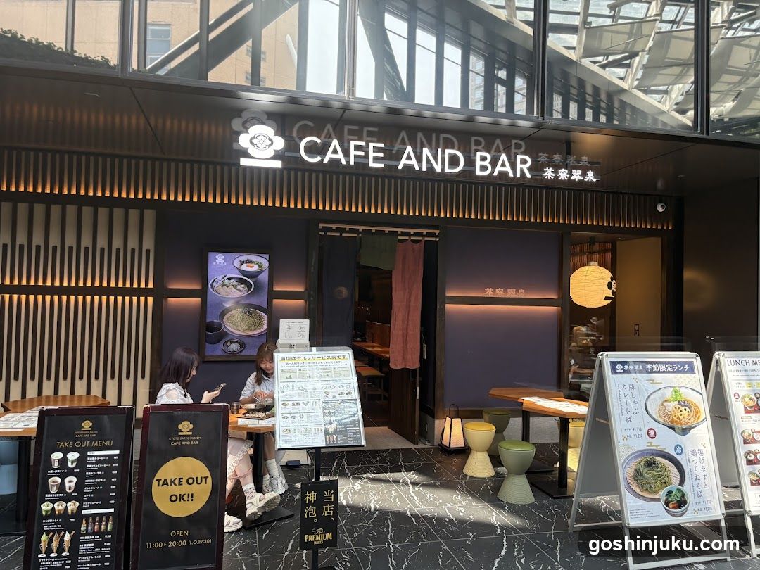 A modern cafe and bar entrance with illuminated signage reading 'CAFE AND BAR' in English and Japanese characters. The facade features vertical wooden slats, a glass door with a pink curtain, and outdoor seating. Menu boards are placed outside, including one highlighting take-out options and another displaying food items like noodles.