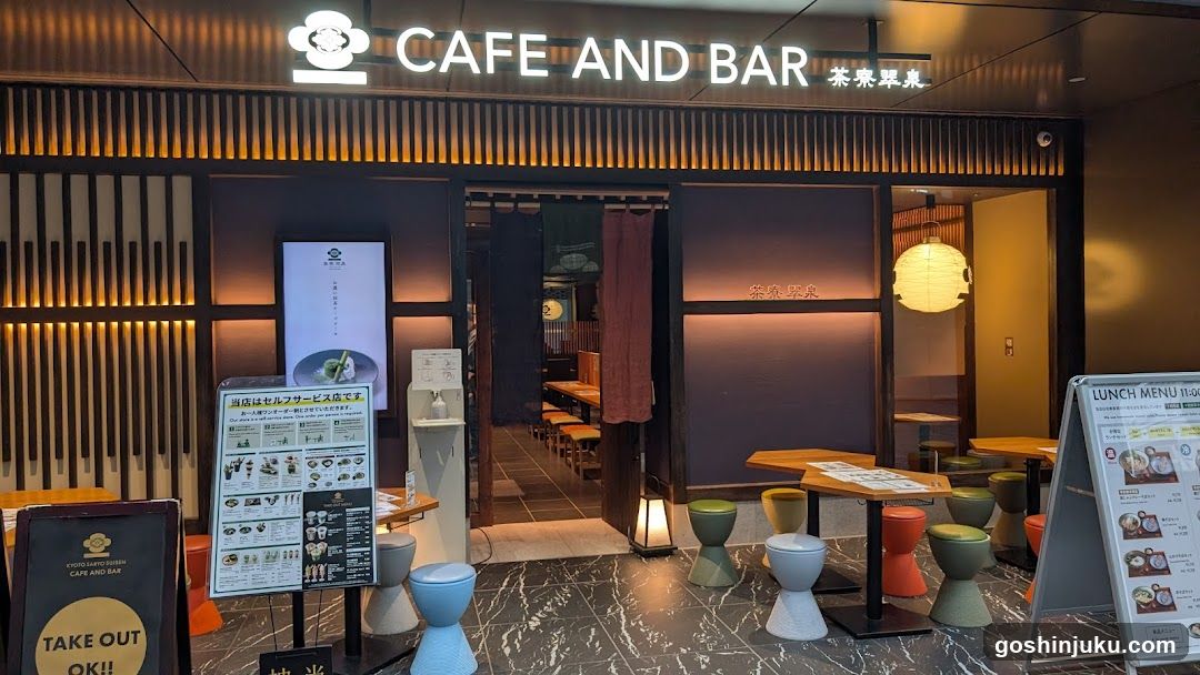 The image shows the storefront of a cafe and bar with warm lighting and wooden accents. The entrance is flanked by two menu boards, one displaying a lunch menu and another showing various food items. There are several stools in different colors arranged outside the entrance.