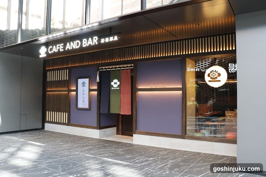 Modern shop front with illuminated signage reading 'CAFE AND BAR' in English and Japanese characters. Wooden slat accents, purple walls, hanging noren curtains (black, green, red), glass windows showing interior seating area, and a circular logo on the window.