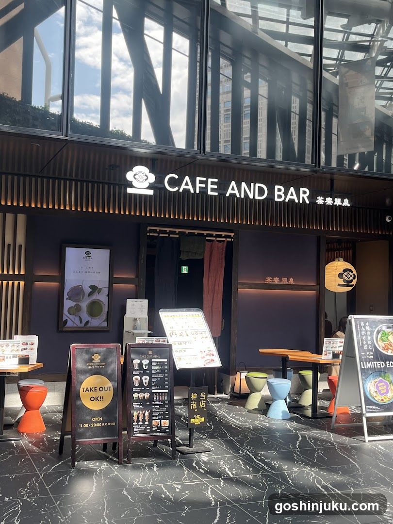A modern cafe and bar storefront with a dark facade, illuminated signage reading 'CAFE AND BAR' in English and Chinese characters. The entrance features glass doors with red curtains. Outside, there are several menu boards displaying food items and prices, along with colorful stools arranged on the tiled pavement. A poster showcasing tea products is visible on the wall.