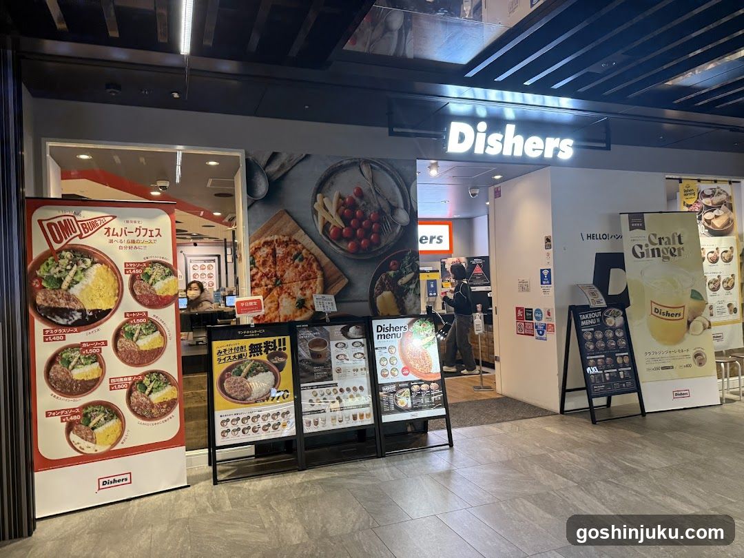 The image shows the entrance of a Dishers restaurant, featuring illuminated signage above the door. Promotional posters display menu items like curry rice and pizza. A staff member is visible inside near the counter. The interior has modern lighting and decor.