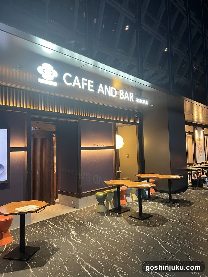 A modern cafe and bar storefront at night, featuring illuminated signage with a chef icon logo. The entrance has glass doors and windows with warm interior lighting visible inside. Outside seating includes wooden tables paired with colorful stools on a marble-patterned floor.
