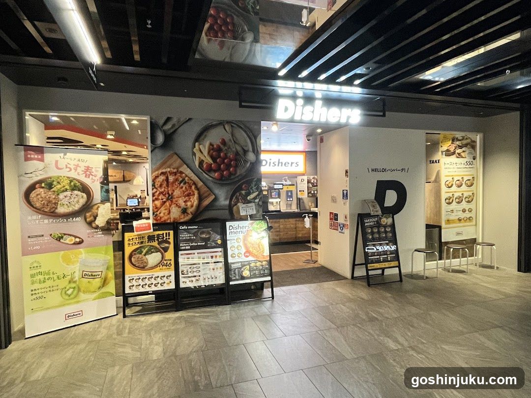 The image shows the entrance of a restaurant called Dishers, with visible signage above and on the wall. There are promotional posters outside displaying food items like pizza and rice bowls. A menu board is placed near the entrance listing various dishes.