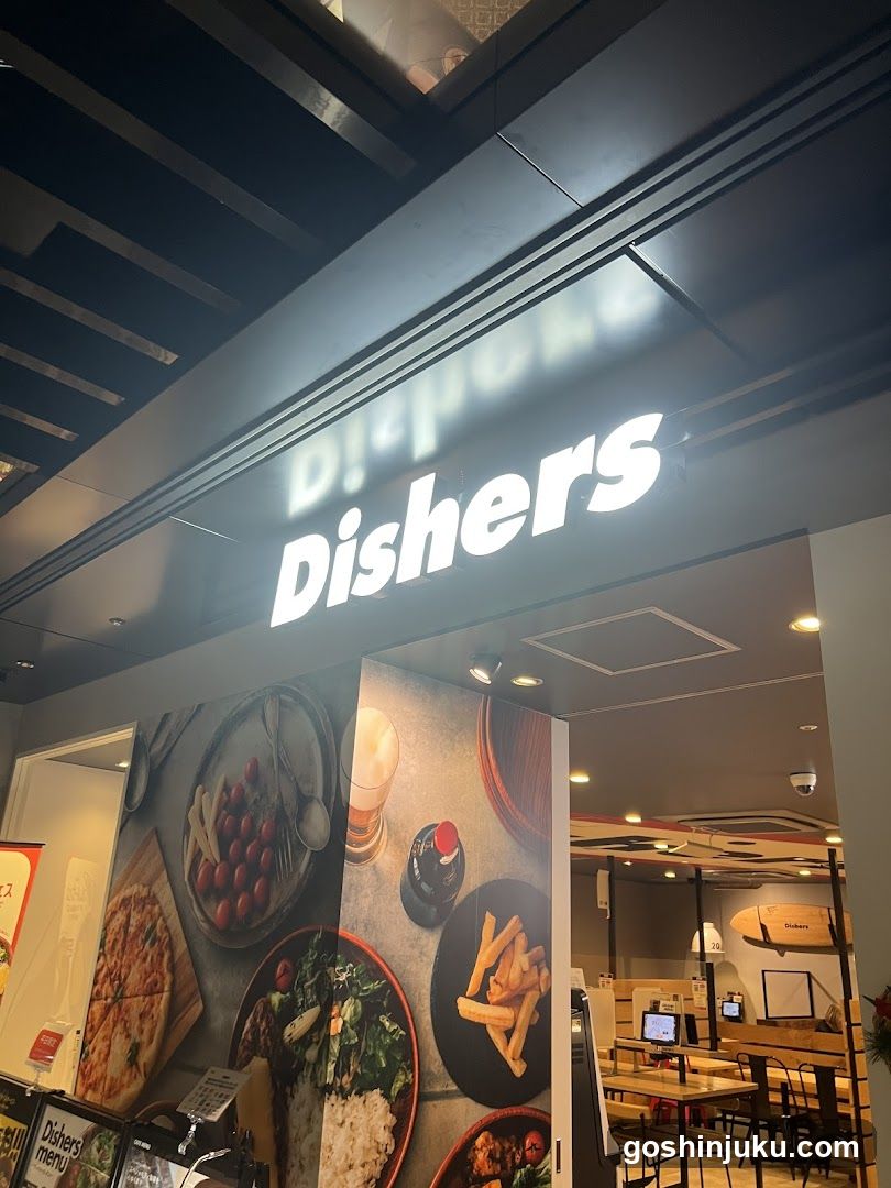 The image shows the exterior entrance of a restaurant named 'Dishers'. The illuminated sign is mounted above the doorway. To the left, there is a large vertical banner displaying images of food items like pizza, salad, and fries. Inside, wooden tables and chairs are visible along with computer monitors on counters.