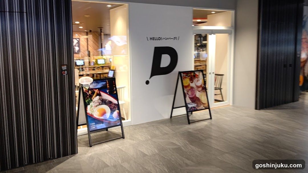 A modern shop entrance with a large 'D' logo and Japanese text on the wall. Two A-frame menu boards display dessert items, including cakes and pastries.