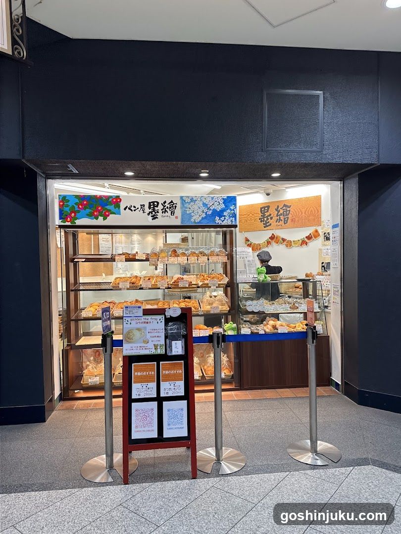 A bakery storefront with glass display cases filled with pastries and breads. The entrance features a signboard with Japanese characters and floral designs, along with a QR code stand at the front.